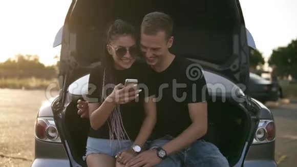 漂亮的年轻的夫妇在一起坐在敞开的汽车后备箱里拥抱着看着手机两者都是视频的预览图