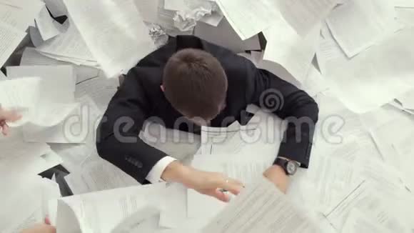 一个疲惫的年轻人坐在一堆文件里签着许多文件办公室职员不能应付工作视频的预览图