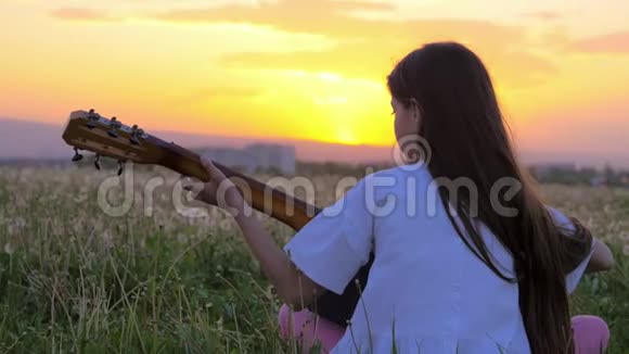 美丽的日落小女孩背着相机坐在蒲公英的田野里弹吉他远离户外视频的预览图