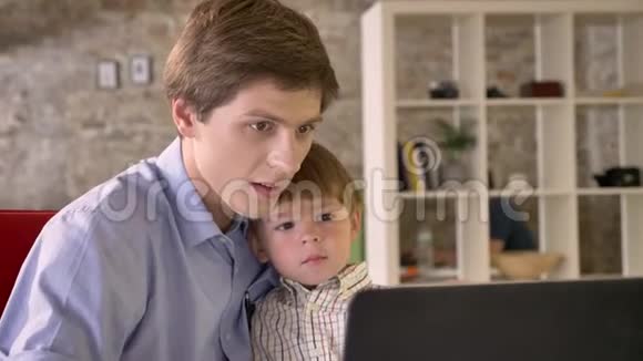 年轻的父亲抱着他微笑的儿子看着笔记本电脑坐在现代的办公室里快乐着视频的预览图