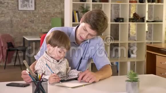 年轻的白种人父亲抱着他的儿子教他如何写作坐在现代的办公室里视频的预览图