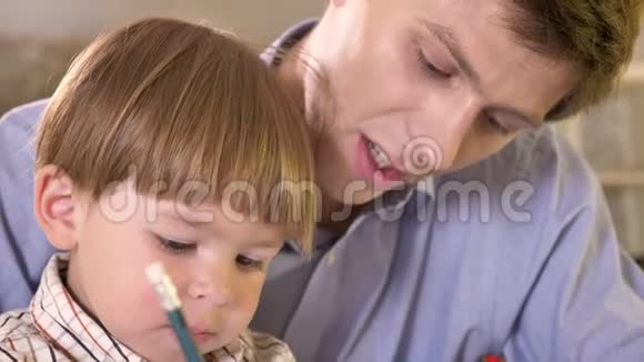 年轻的父亲和他的小儿子在纸上画画拿着平板电脑爸爸帮助和教他的儿子如何写作视频的预览图