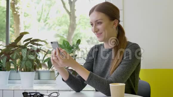 扎着马尾辫的年轻美女坐在咖啡馆里用电话向前看微笑着视频的预览图