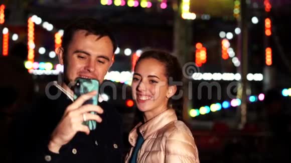 年轻漂亮的情侣在一起约会晚上在游乐园用智能手机自拍视频的预览图