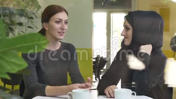 两个年轻漂亮的女人坐在咖啡馆里其中一个是穆斯林女人戴着头巾说话微笑从街上看视频的预览图