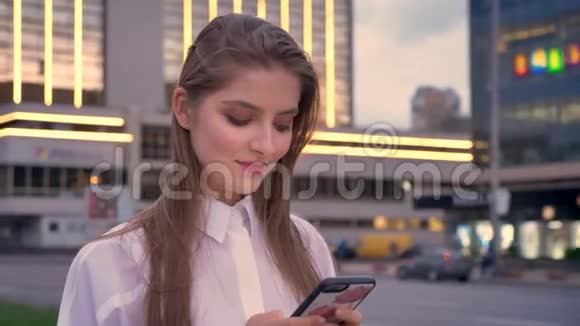 年轻迷人的精力充沛的女孩正在夏季市中心的日落时分在智能手机上输入信息视频的预览图