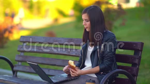 一个迷人的年轻女孩吃一个面包在笔记本电脑上工作视频的预览图
