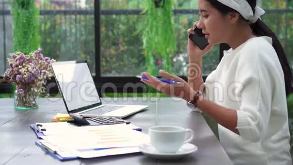 美丽的年轻压力亚洲妇女在笔记本电脑上工作而不高兴使用智能手机在客厅在家里视频的预览图