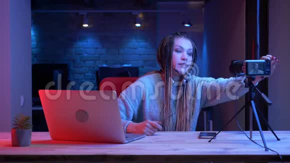 使用带有霓虹灯背景的笔记本电脑拍摄的耳机中戴着可怕锁的年轻迷人女性博主的特写镜头视频的预览图