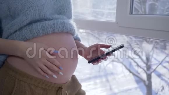 准妈妈一边摸着她赤裸的肚子一边用手机一边站在一扇大窗户一边戴着手套视频的预览图