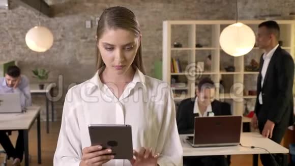 年轻集中精力的女商人正在办公室里敲打平板电脑看着镜头她的同事们正在与视频的预览图