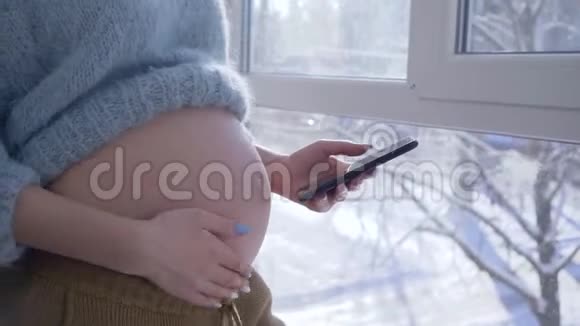 怀孕的女孩站在公寓的一扇大窗户旁用手机抚摸着她赤裸的肚子视频的预览图