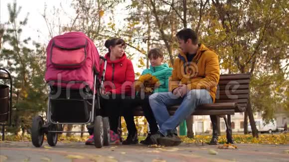 爸爸妈妈带着孩子坐在秋天公园里的长椅上带着婴儿车聊天视频的预览图