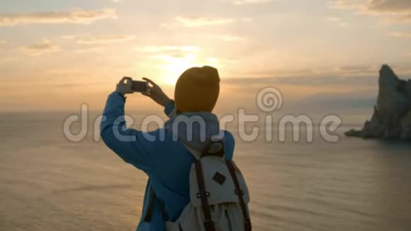 年轻迷人的女孩徒步旅行在智能手机上拍照在海边的海角上拍摄美丽的日落达到了目标视频的预览图