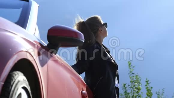 一个穿着皮夹克和太阳镜的年轻时髦黑发女孩站在一辆红色敞篷车旁边视频的预览图