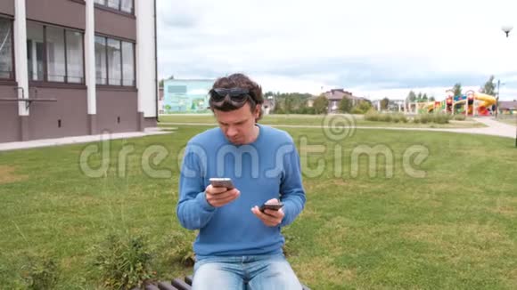 男人拿着两部手机坐在家附近的长凳上调用和键入消息视频的预览图