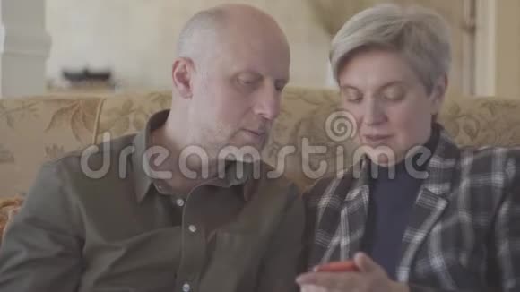 一对成年夫妇坐在大沙发上秃头的女人在手机上为他的灰短老头拍照视频的预览图