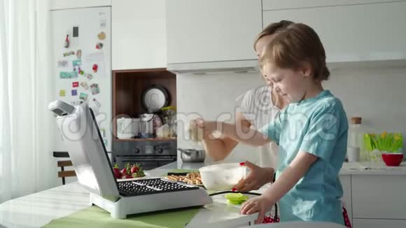 年轻的女人在厨房做华夫饼可爱的男孩帮助她一大早就不规律地做早餐视频的预览图