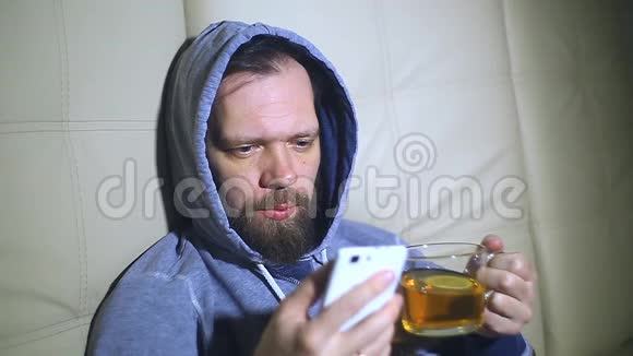大胡子男人在智能手机屏幕上看新闻喝茶视频的预览图