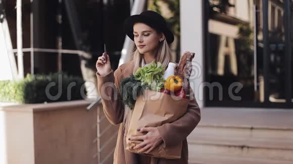 年轻漂亮的女人穿着时髦的外套站在街上拿着一包产品拿着信用卡视频的预览图