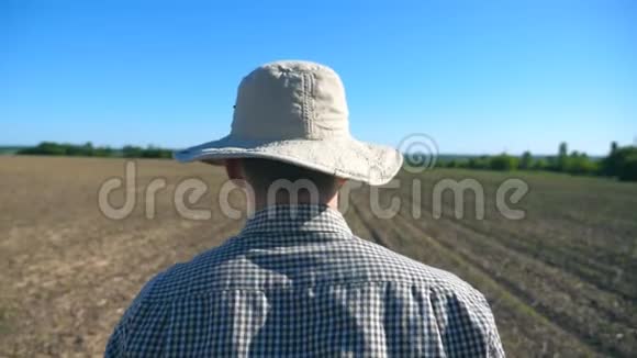 在阳光明媚的夏日跟随着无法辨认的戴着帽子和衬衫的年轻男性农民在他的田野上散步蓝天视频的预览图