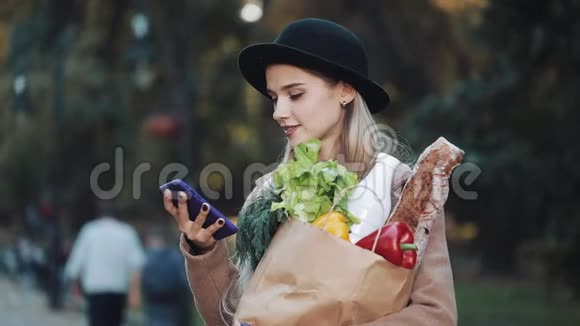 年轻漂亮的女人穿着时髦的外套站在公园里拿着一包产品用智能手机说话视频的预览图