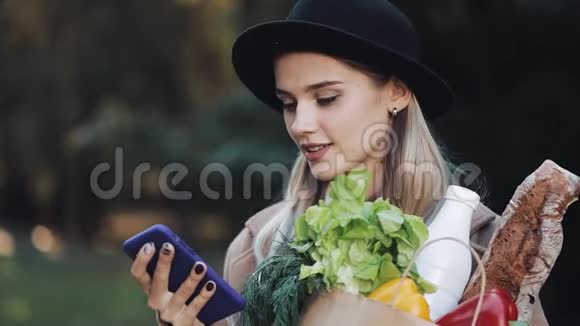年轻漂亮的女人穿着时髦的外套站在公园里拿着一包产品用智能手机说话视频的预览图
