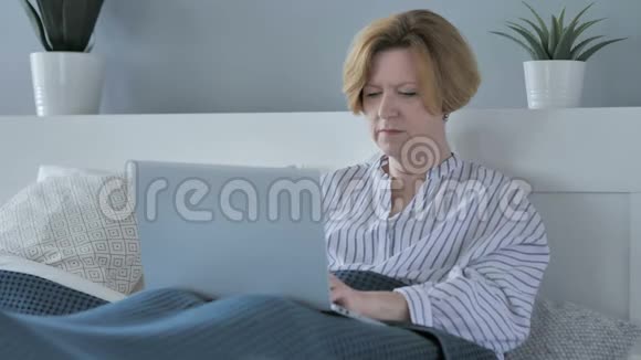 快乐高年级女性在床上使用笔记本电脑的成功反应视频的预览图