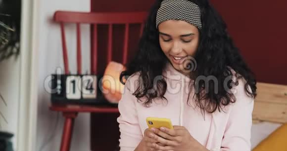 漂亮的年轻女人一边微笑一边在黄色的智能手机上聊天坐在家里的床上女孩向视频的预览图