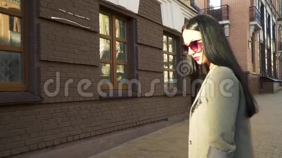时尚的年轻女性戴着太阳镜在街道上的棕色建筑上穿外套视频的预览图