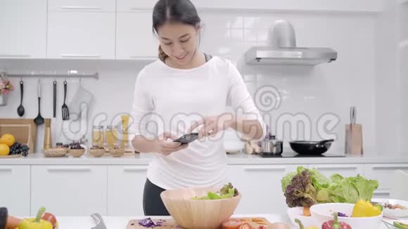 年轻的亚洲博客女性在厨房的社交媒体上使用智能手机照片帖子女性在家做沙拉视频的预览图