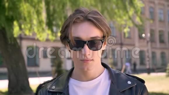 年轻的帅哥穿着黑色皮夹克举起眼镜看着相机站在城市的街道上视频的预览图