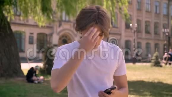 年轻的帅哥穿着白色衬衫打电话站在大学附近的公园里男学生打电话视频的预览图