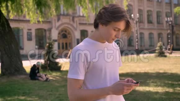 年轻的帅哥穿着白色衬衫在智能手机上打字看着相机学生站在大学附近的公园里视频的预览图