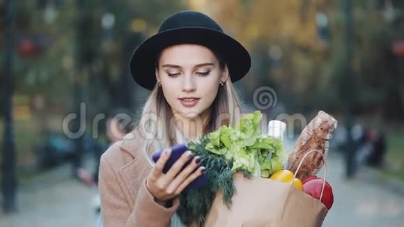 年轻漂亮的女人穿着时髦的外套站在公园里拿着一包产品用智能手机说话视频的预览图