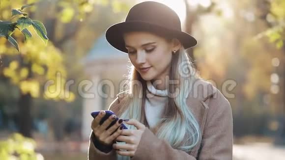 年轻女子穿着外套用智能手机站在秋天公园女孩看着镜头户外技术视频的预览图