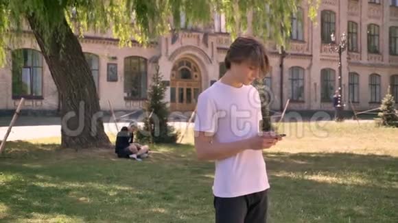 年轻漂亮的男学生穿着白色衬衫站着拿着电话公园靠近大学背景视频的预览图
