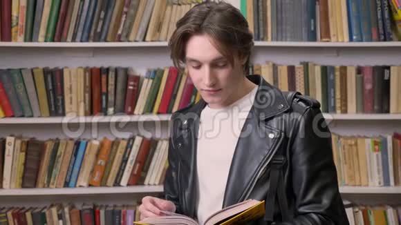 年轻的帅哥穿着黑色皮夹克在图书馆看书微笑着正在学习的男学生的画像视频的预览图