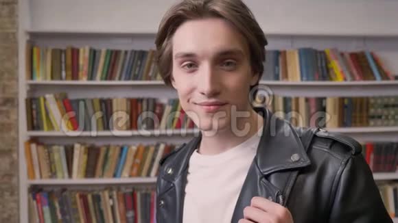 年轻的帅哥穿着黑色皮夹克站在图书馆里看着相机微笑着书架背景视频的预览图