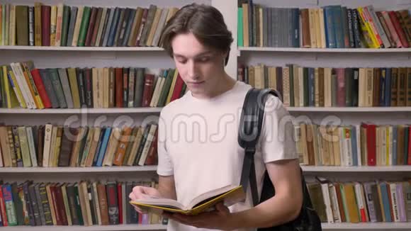 年轻的美男子穿着白衬衫站在图书馆看书看着相机拿着书书架视频的预览图