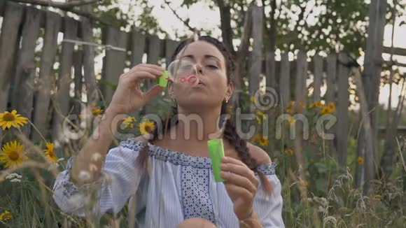 特写镜头美丽的女人让肥皂泡坐在靠近木制围栏的草地上视频的预览图