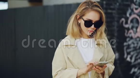 美丽的金发女郎正在使用智能手机触摸屏幕站在户外背景墙上涂鸦年轻女子视频的预览图