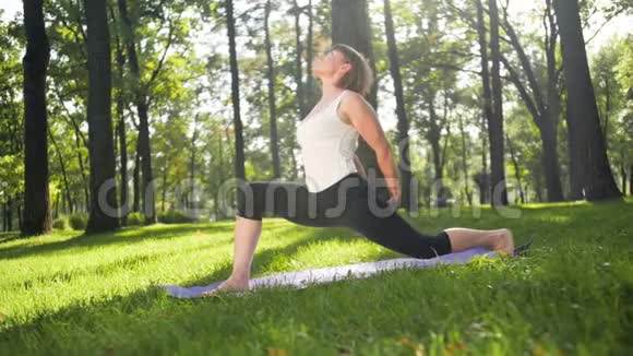 在公园的健身垫上伸展快乐微笑的女人的4K镜头中年妇女在森林做瑜伽练习视频的预览图