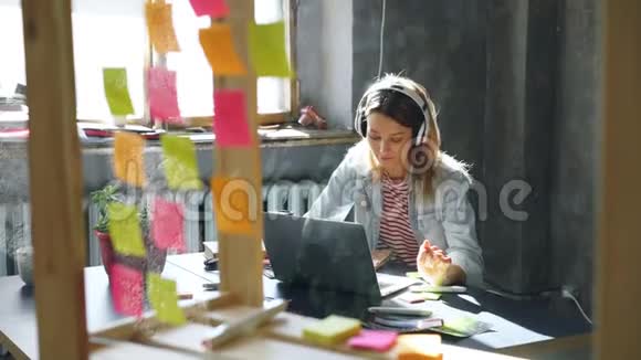漂亮的金发女孩正在听音乐在漂亮的阁楼办公室里用笔记本电脑工作她在记事本上做笔记视频的预览图