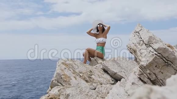 穿着泳衣太阳镜和草帽的漂亮黑发女人坐在背景蓝色大海上的岩石上视频的预览图