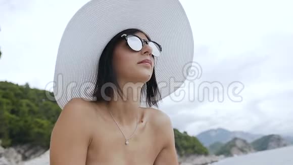 一位身穿白色泳衣戴着大帽子和太阳镜的年轻女子坐在岩石海滩上享受阳光和水夏天视频的预览图