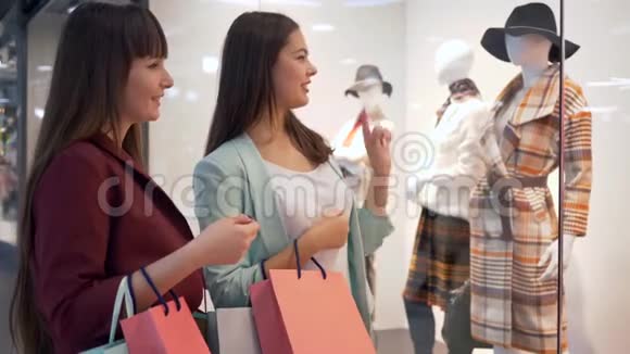 年轻的女朋友带着袋子站在商店橱窗附近收集新的衣服并讨论在商场购物视频的预览图