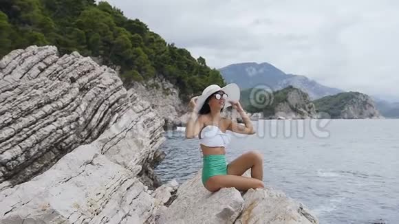 年轻漂亮的女人穿着泳衣戴着宽帽子戴着太阳镜坐在海边的岩石上暑假假期视频的预览图