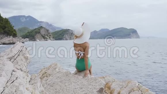 美丽的黑发女孩戴着夏草帽和泳衣的肖像她坐在亚得里亚海旁边的岩石上视频的预览图