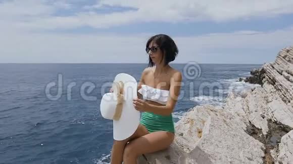 戴墨镜的漂亮女孩坐在岩石上穿着泳衣戴着海蓝色海水背景的帽子视频的预览图